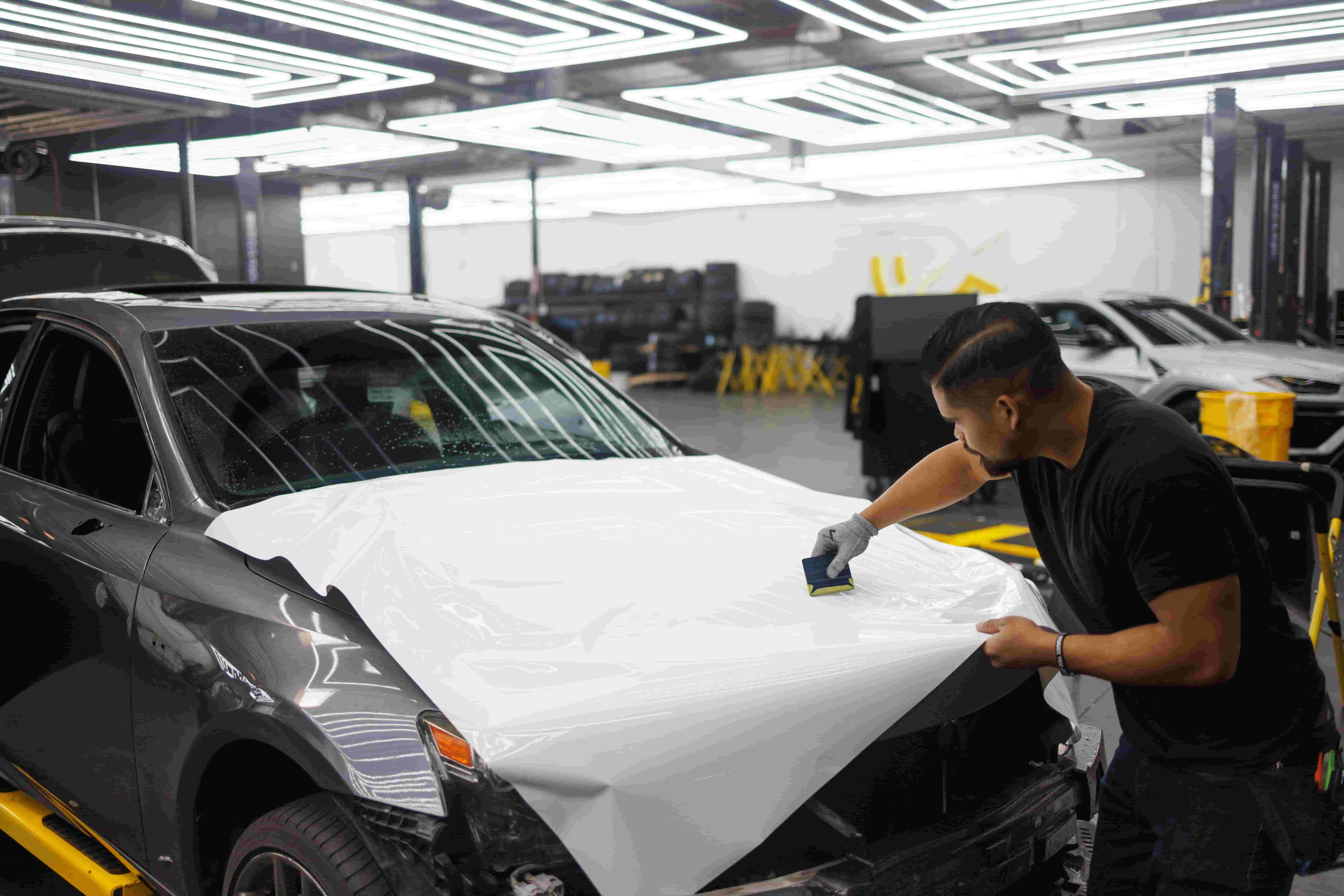 Man applying paint protection film to a car hood in a workshop setting.