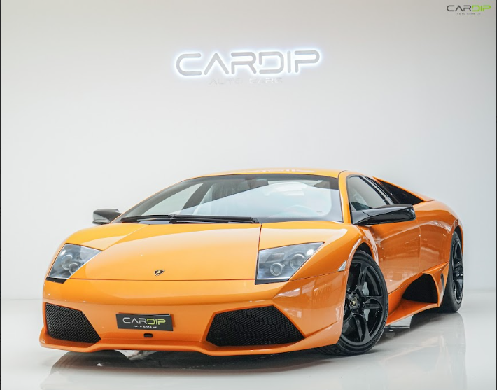 An orange Lamborghini sports car displayed in a clean, white showroom with a glossy floor and a glowing “CARDIP Auto Care, LLC” sign on the wall behind it.
